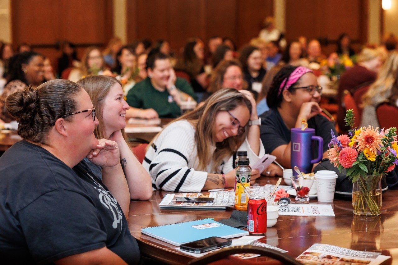 A woman takes notes at a table, smiling, while other early child care professionals around her watch a speaker.