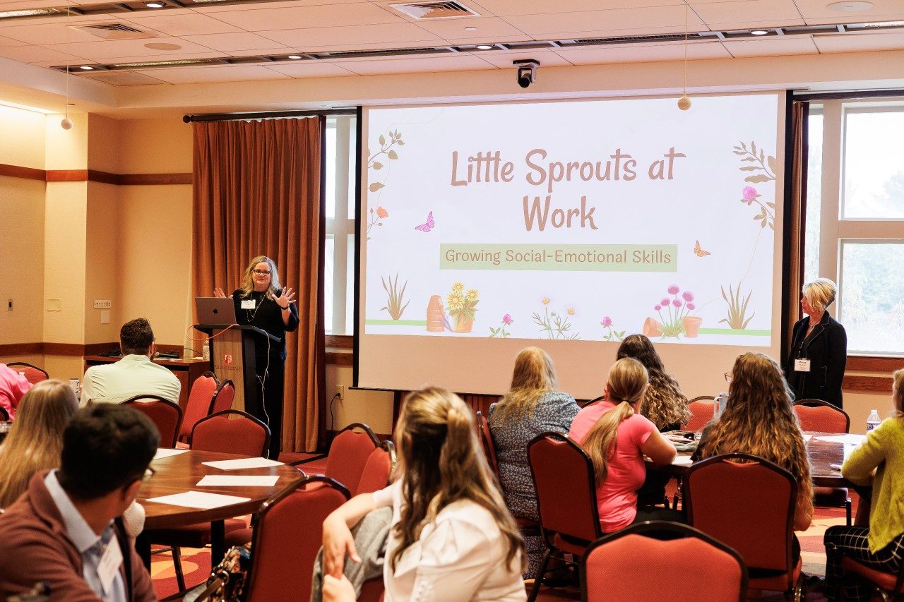 Two presenters stand at the front of a class of teachers with a powerpoint that says "little sprouts at work, growing social-emotional skills".