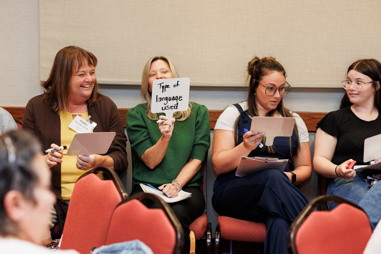 Child care professionals sit along a wall and write on white board paddles, with one holding up a board saying "type of language used".