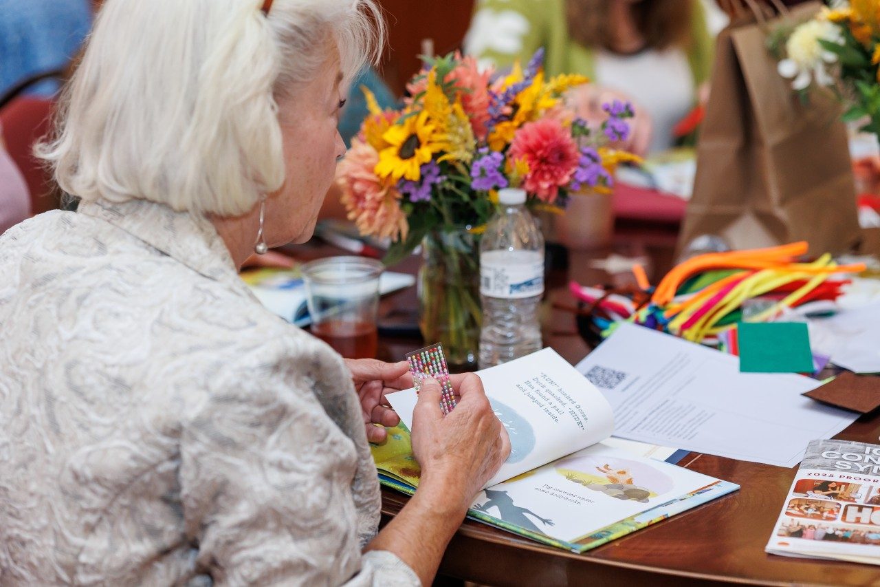 A child care professional sits at a conference table with flowers and arts & crafts supplies, adding stickers to a book.